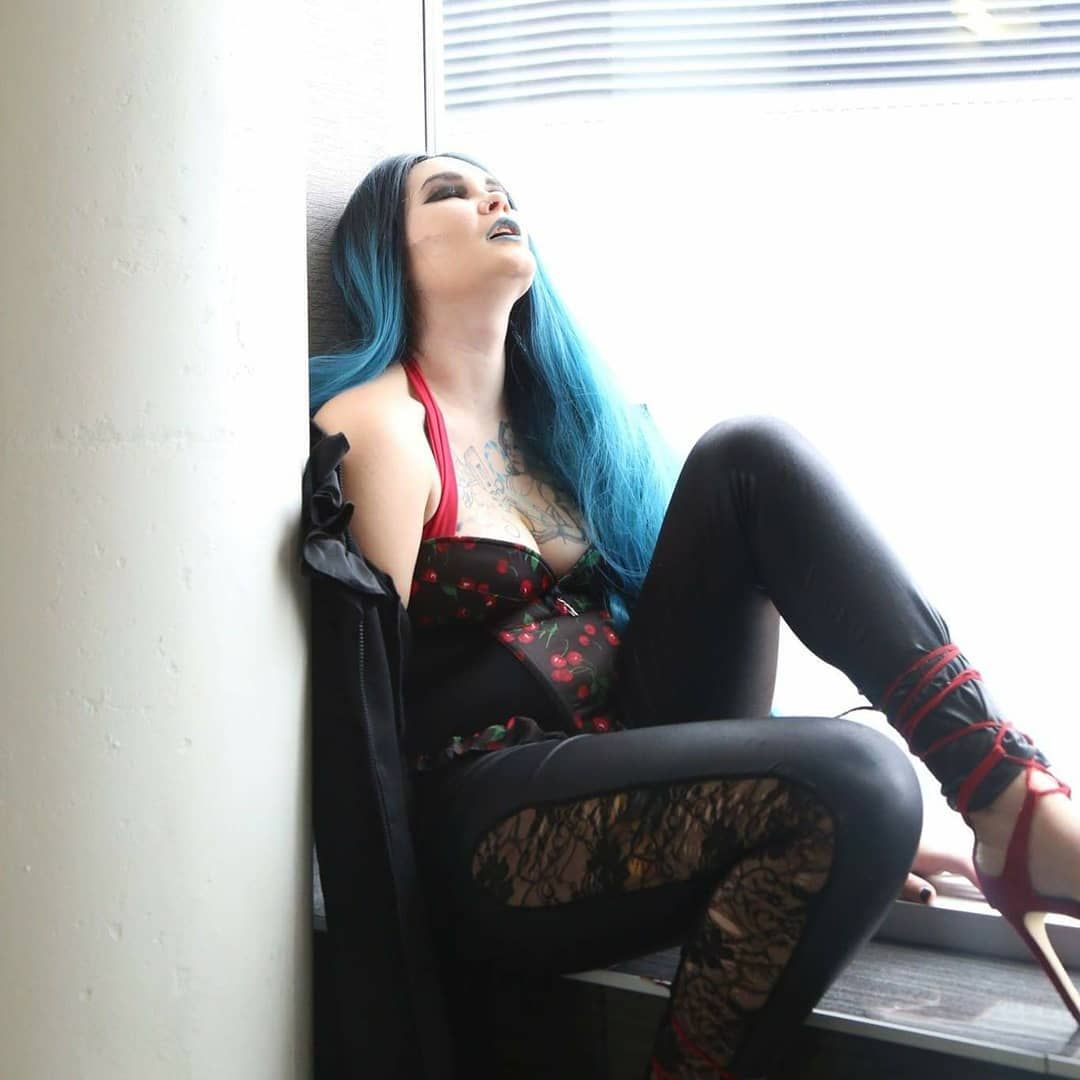 Woman with long blue hair in gothic outfit sitting against wall by window in black pants and floral corset.