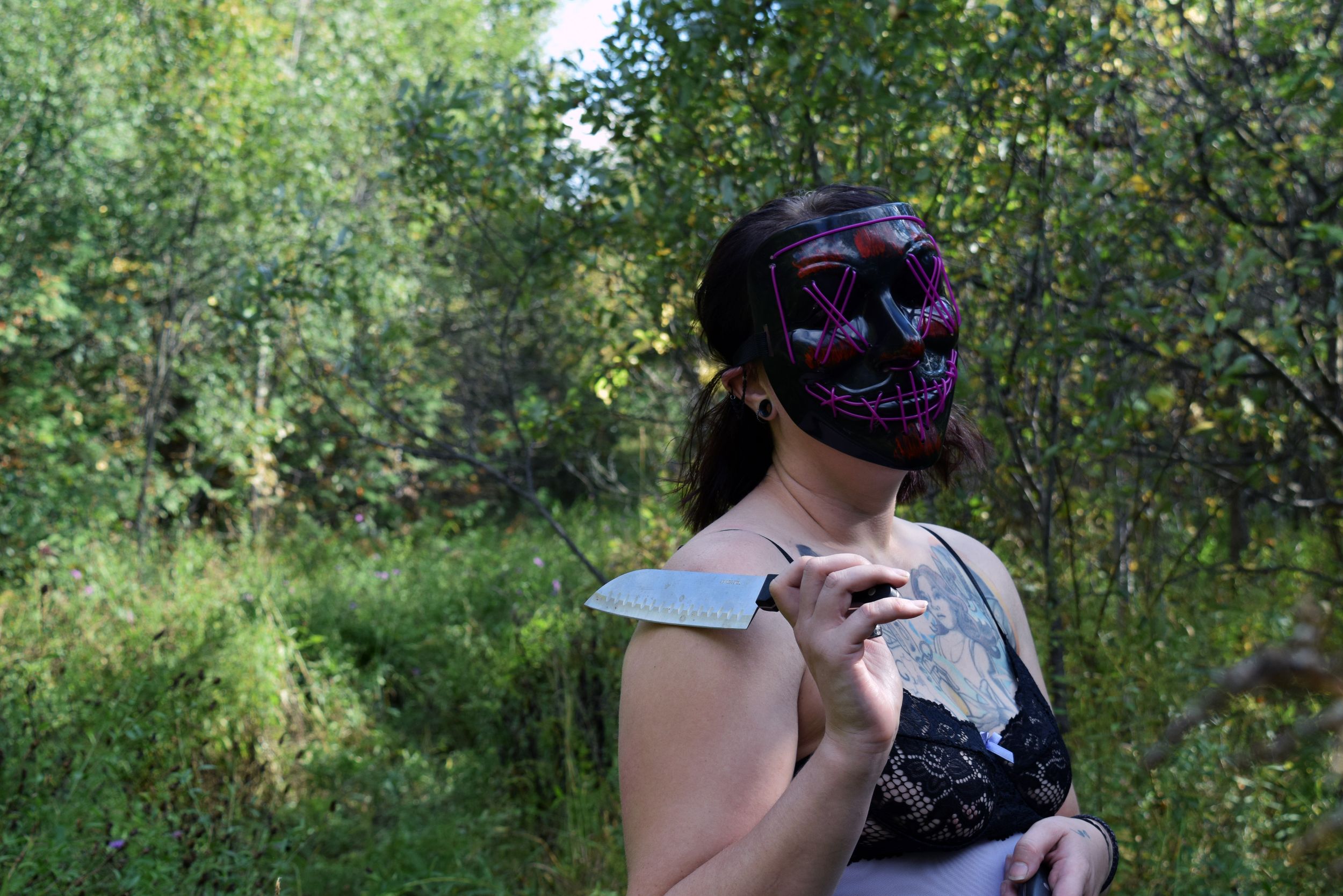 Woman wearing glowing purple skull mask holding a knife outdoors in a forest setting.