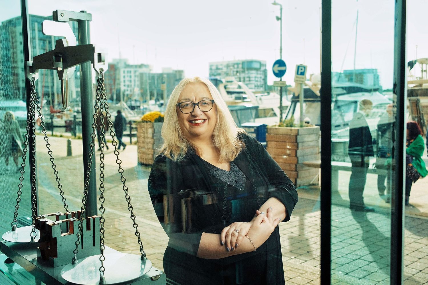 Woman in black outfit smiling near waterfront with urban buildings in background.