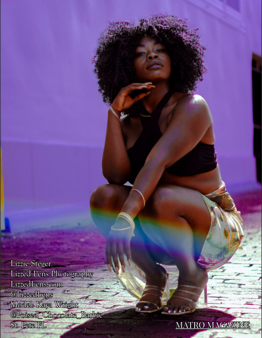 A woman with natural curly hair crouches gracefully on a colorful street, wearing a black crop top and rainbow-light pants.