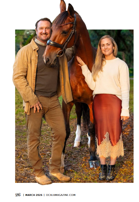 A smiling couple stands with a bay horse outdoors, woman in cream sweater and rust skirt, man in tan jacket and khakis.