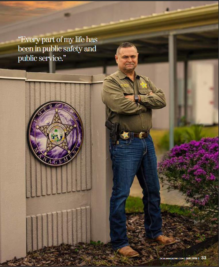 Law enforcement officer in uniform stands beside a sheriff's office sign, arms crossed, with purple flowers in background.