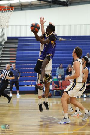 Basketball player in blue jersey leaps high for a shot against defenders in white uniforms during indoor game.