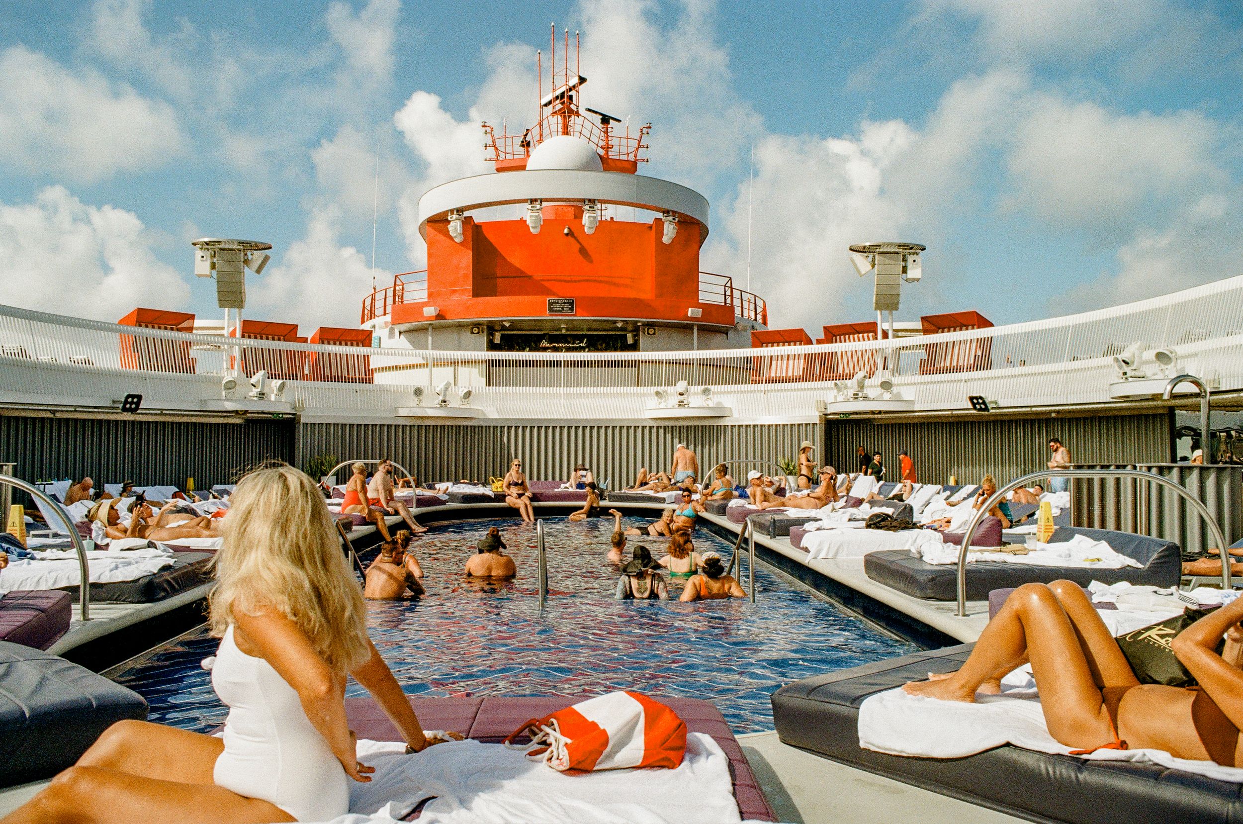 Passengers relaxing by pool deck on cruise ship with distinctive orange and white structure.