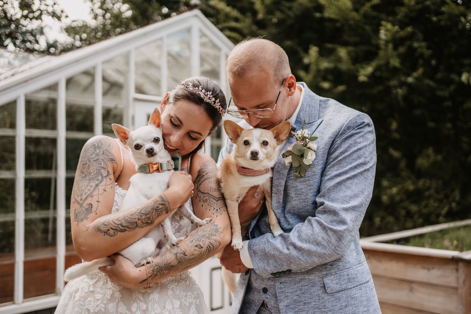 Newlyweds pose with their two small white dogs in front of a greenhouse.