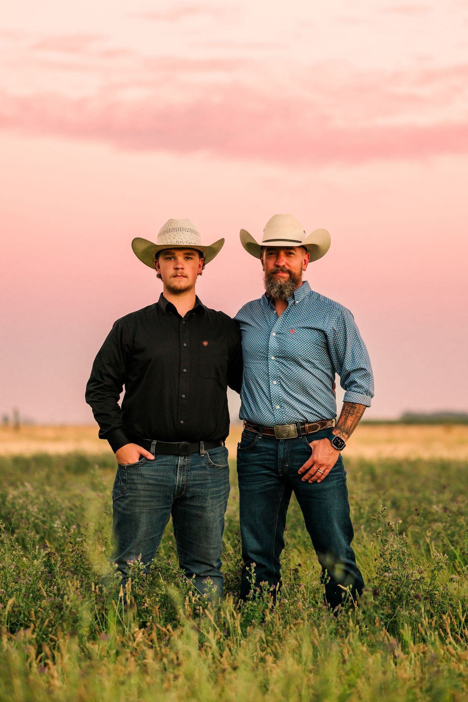 Two people wearing cowboy hats and western attire pose in field during sunset.