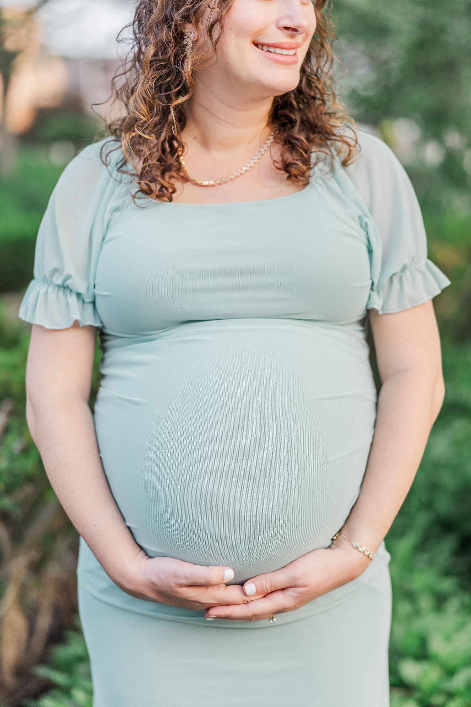 Woman in mint green dress with flutter sleeves shows off baby bump in garden maternity photos.