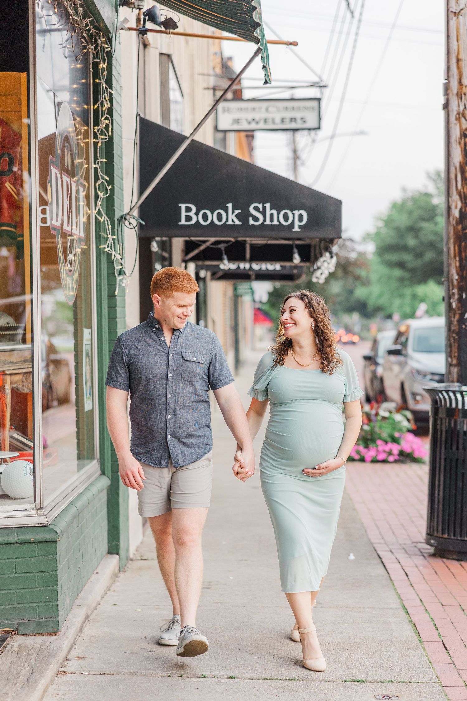 A couple strolls along a brick sidewalk past storefronts during their maternity session.