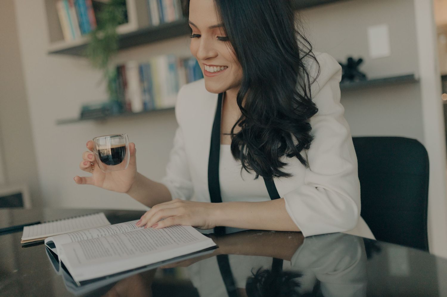 Professional in white blazer reading while enjoying coffee at desk with books.