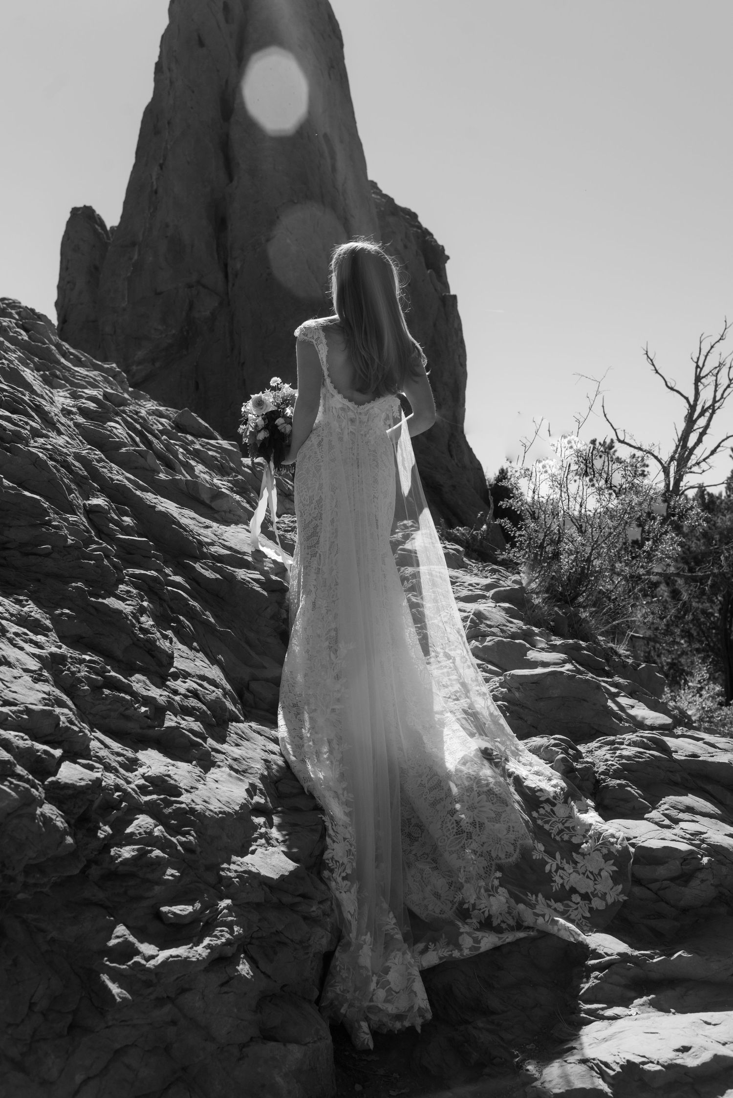 Black and white photo of someone in a flowing white dress standing on rocky terrain against a bright sun in the sky.