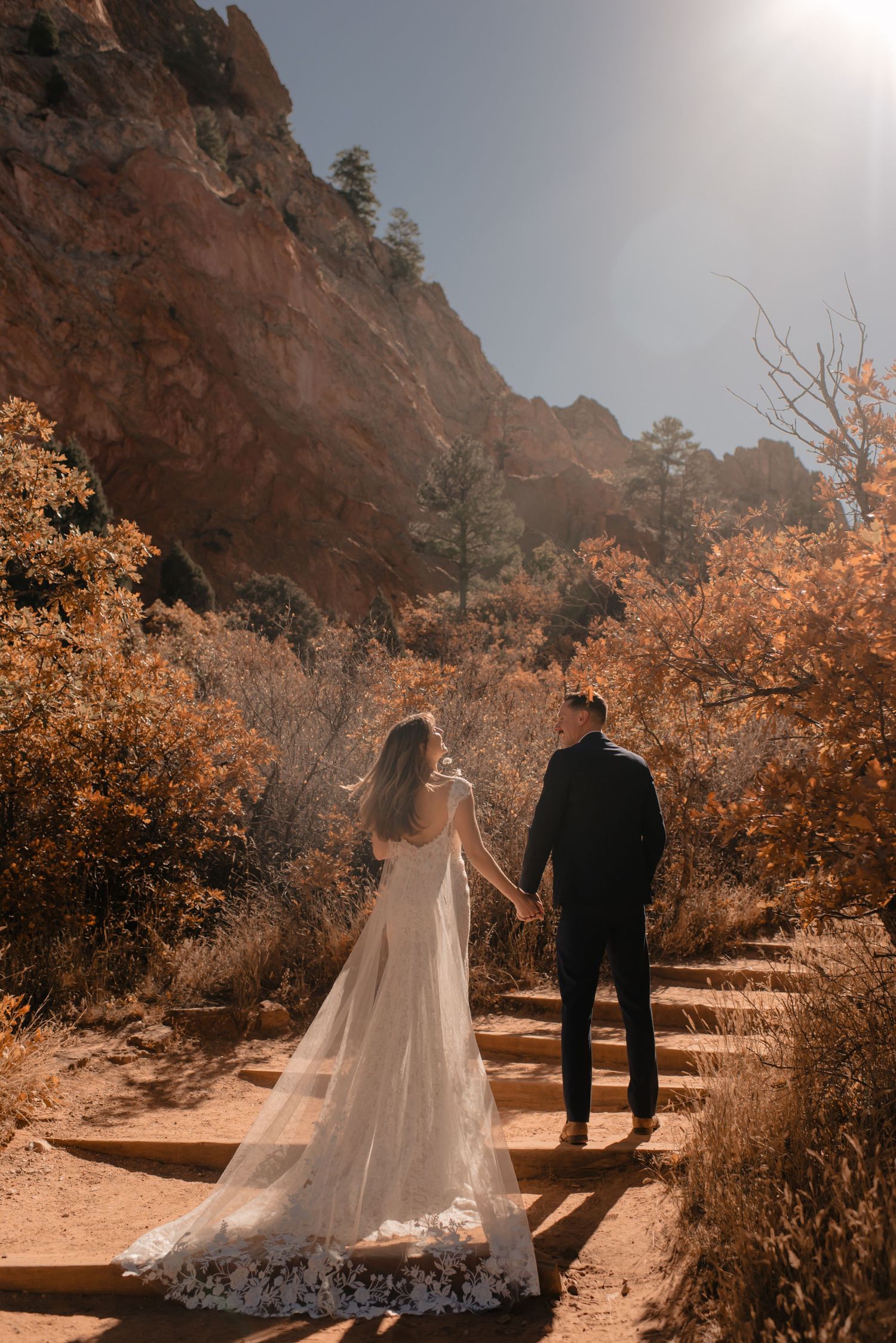 A couple in wedding attire stand on a mountain trail surrounded by autumn foliage and red rock cliffs.