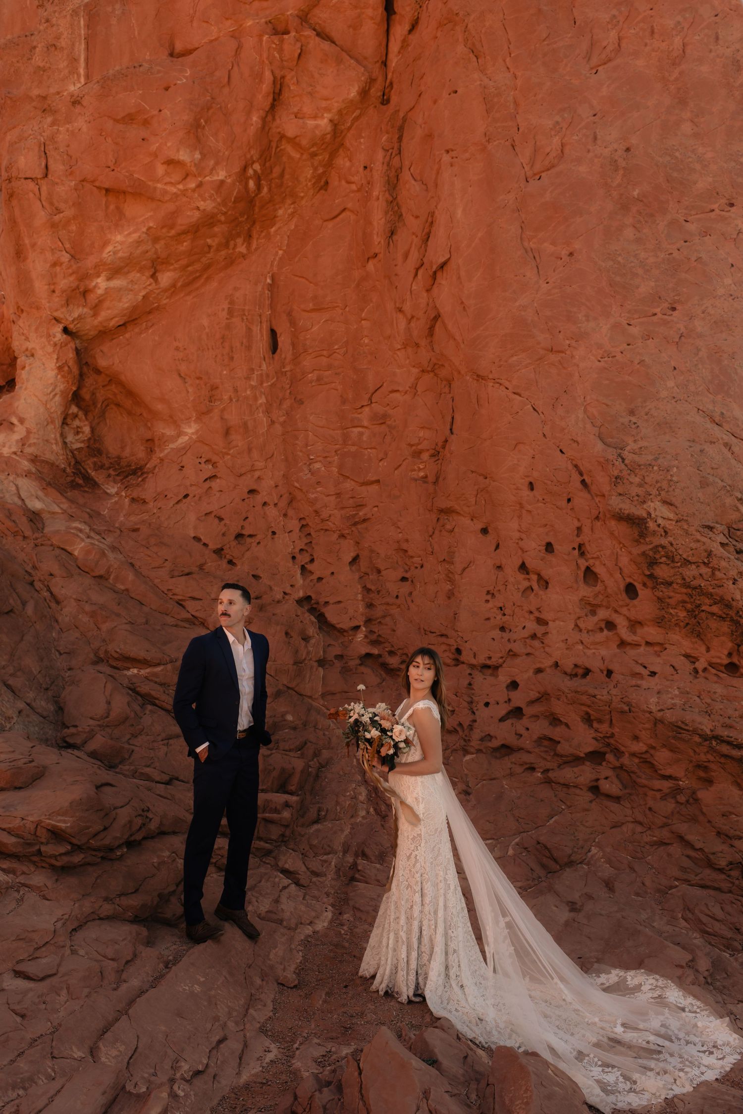 A wedding couple poses against dramatic red rock formations in a desert landscape.