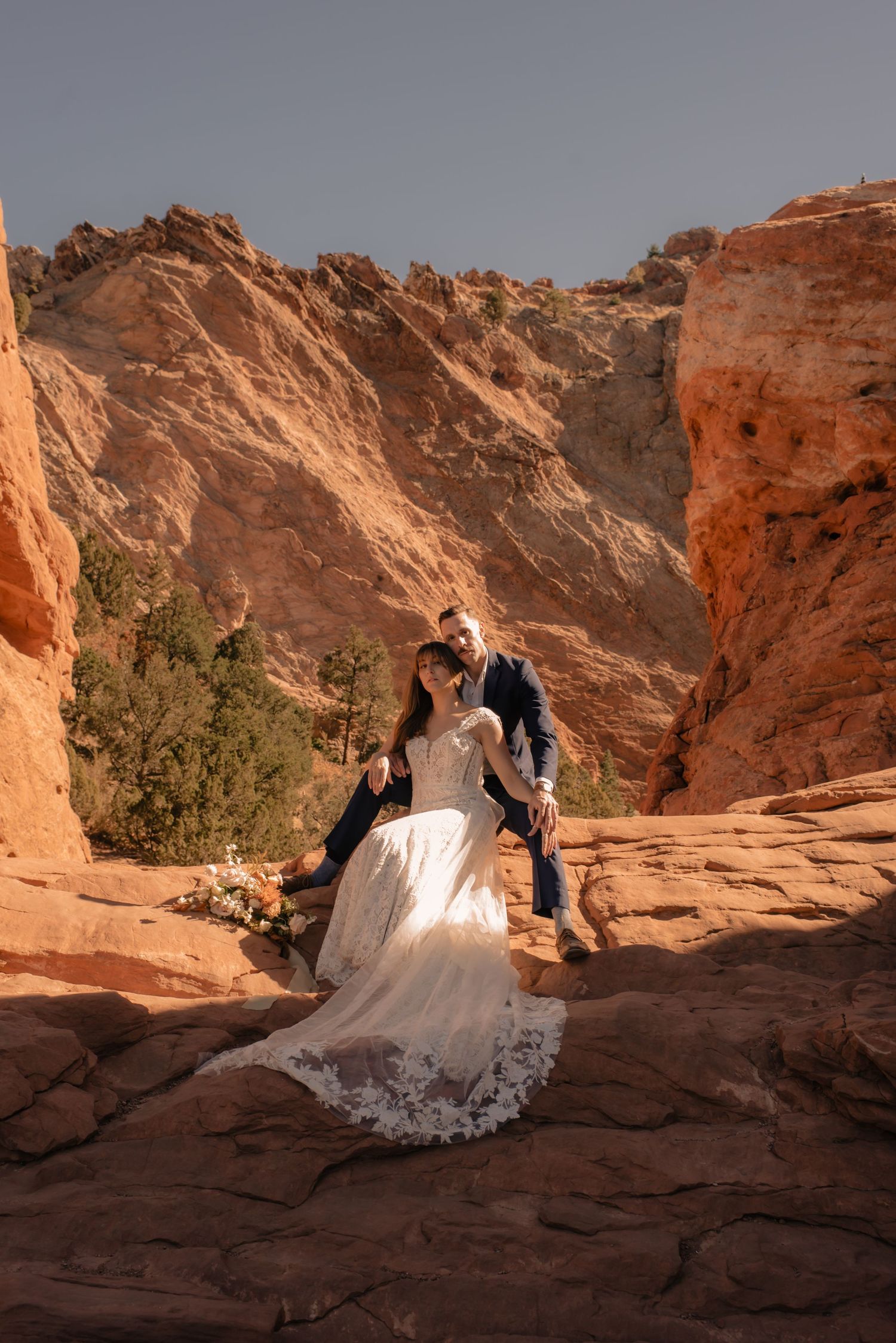 A wedding couple share an intimate moment on red sandstone rocks in a dramatic desert landscape.