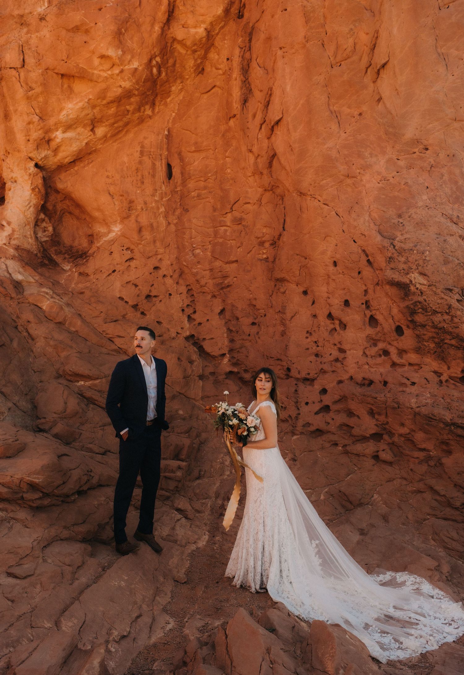 Wedding couple poses against dramatic red rock formation in desert landscape with flowing white dress and bouquet.