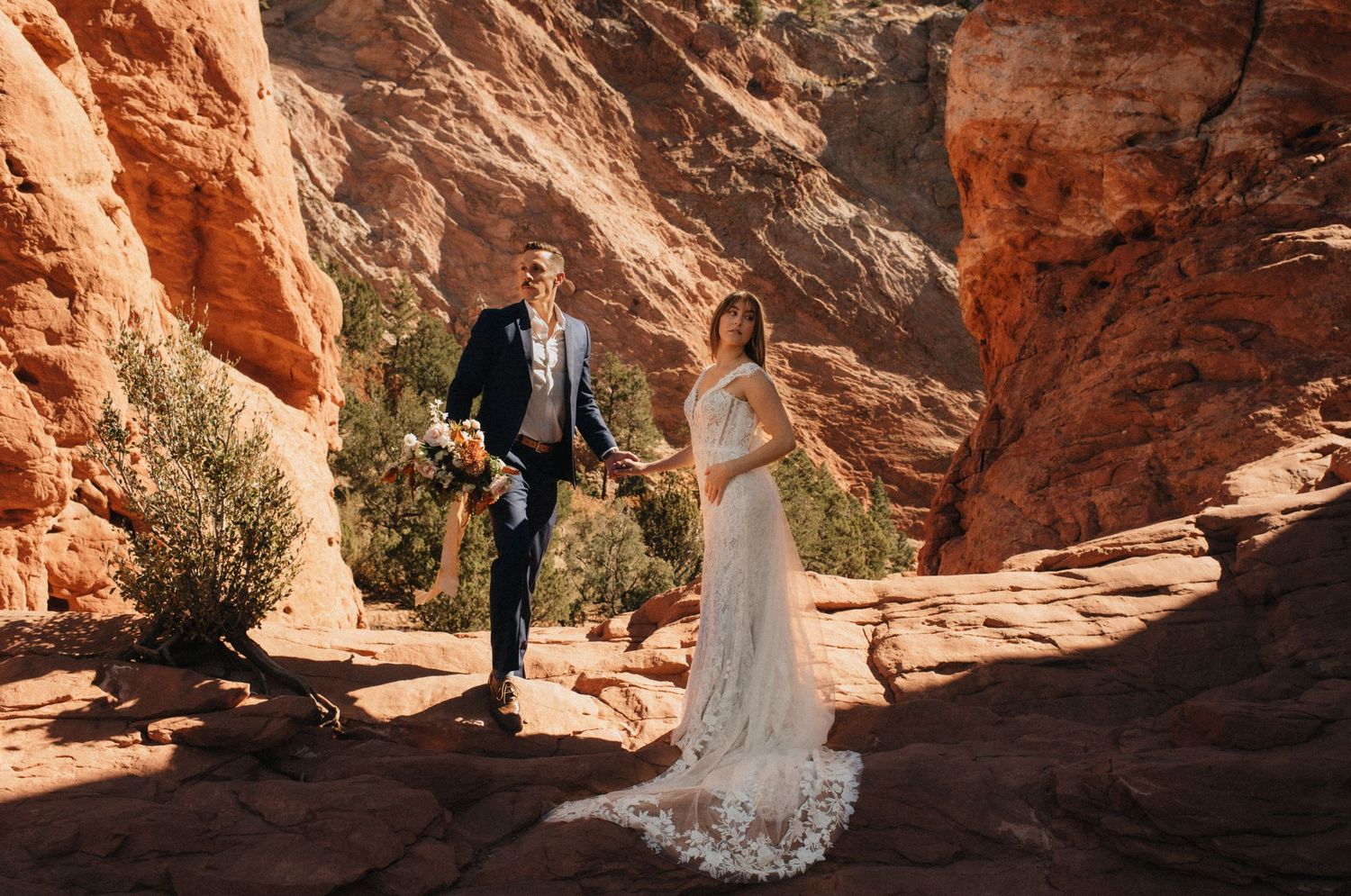 A couple in wedding attire walk together through stunning red rock formations in a desert landscape.
