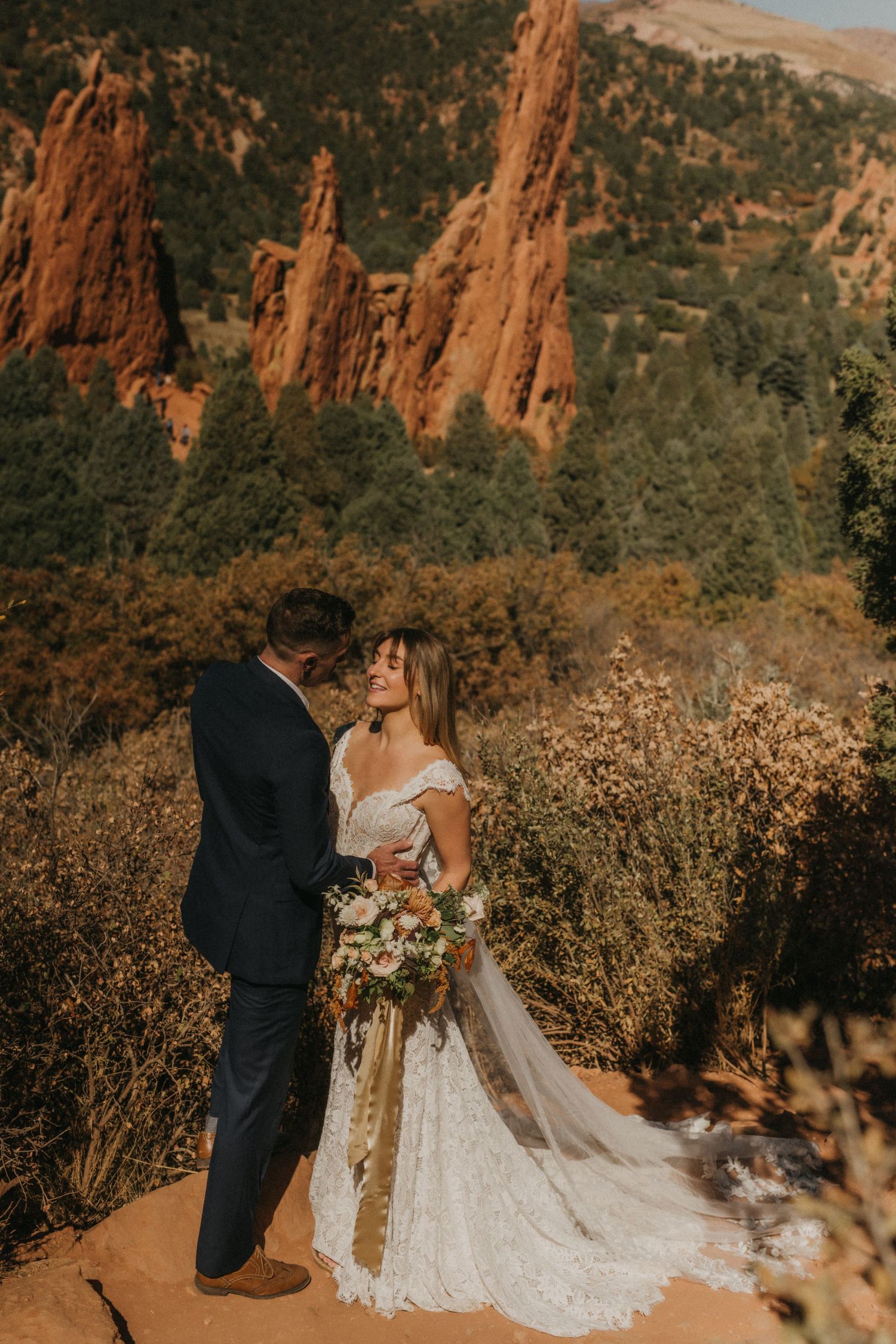 A couple shares an intimate moment in front of towering red rock formations at sunset in a desert landscape.