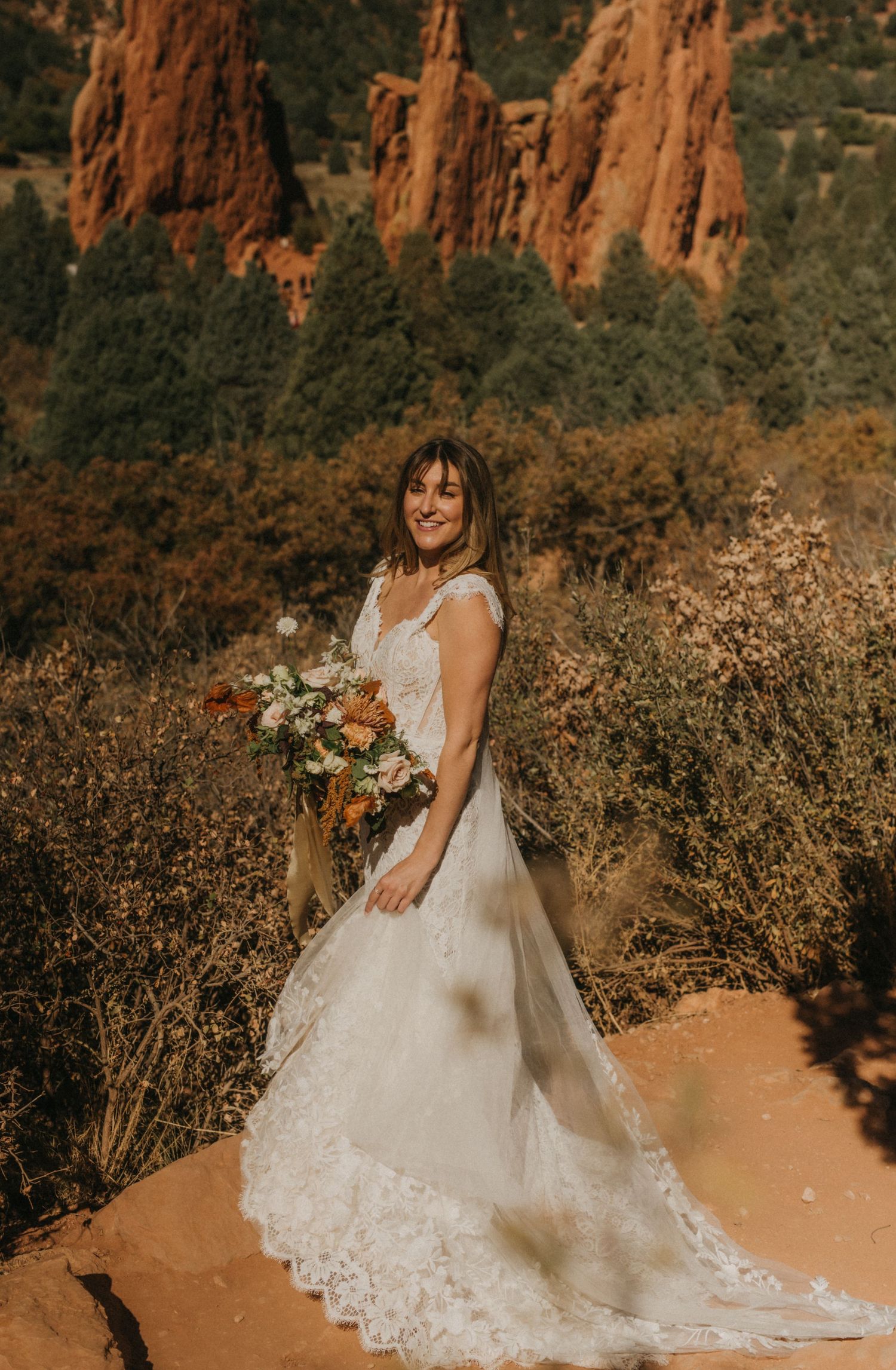A white lace wedding dress flows elegantly against the backdrop of majestic red rock cliffs and desert brush.
