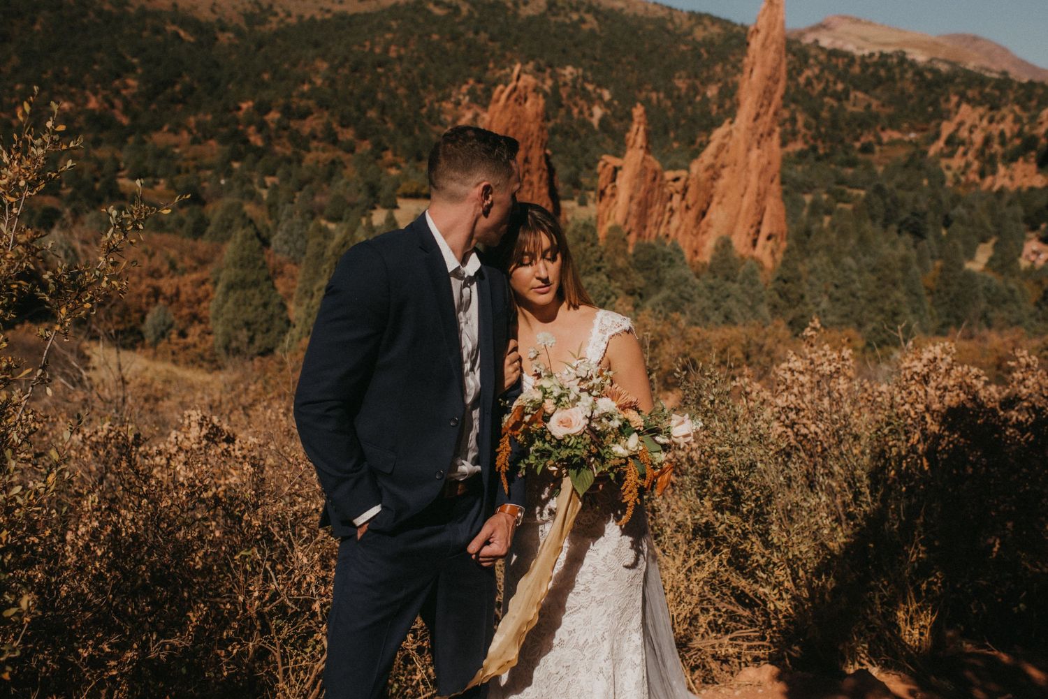 A couple in wedding attire stands together with red rock formations at Garden of the Gods mountain backdrop.