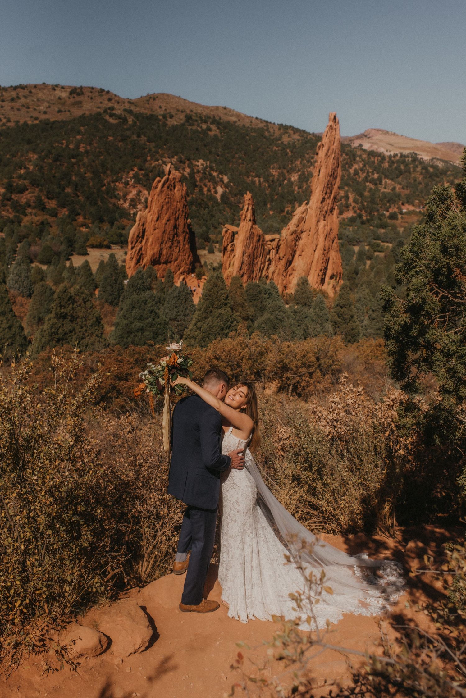 Newlyweds embrace on a dirt path with towering orange rock spires and mountains in the background.