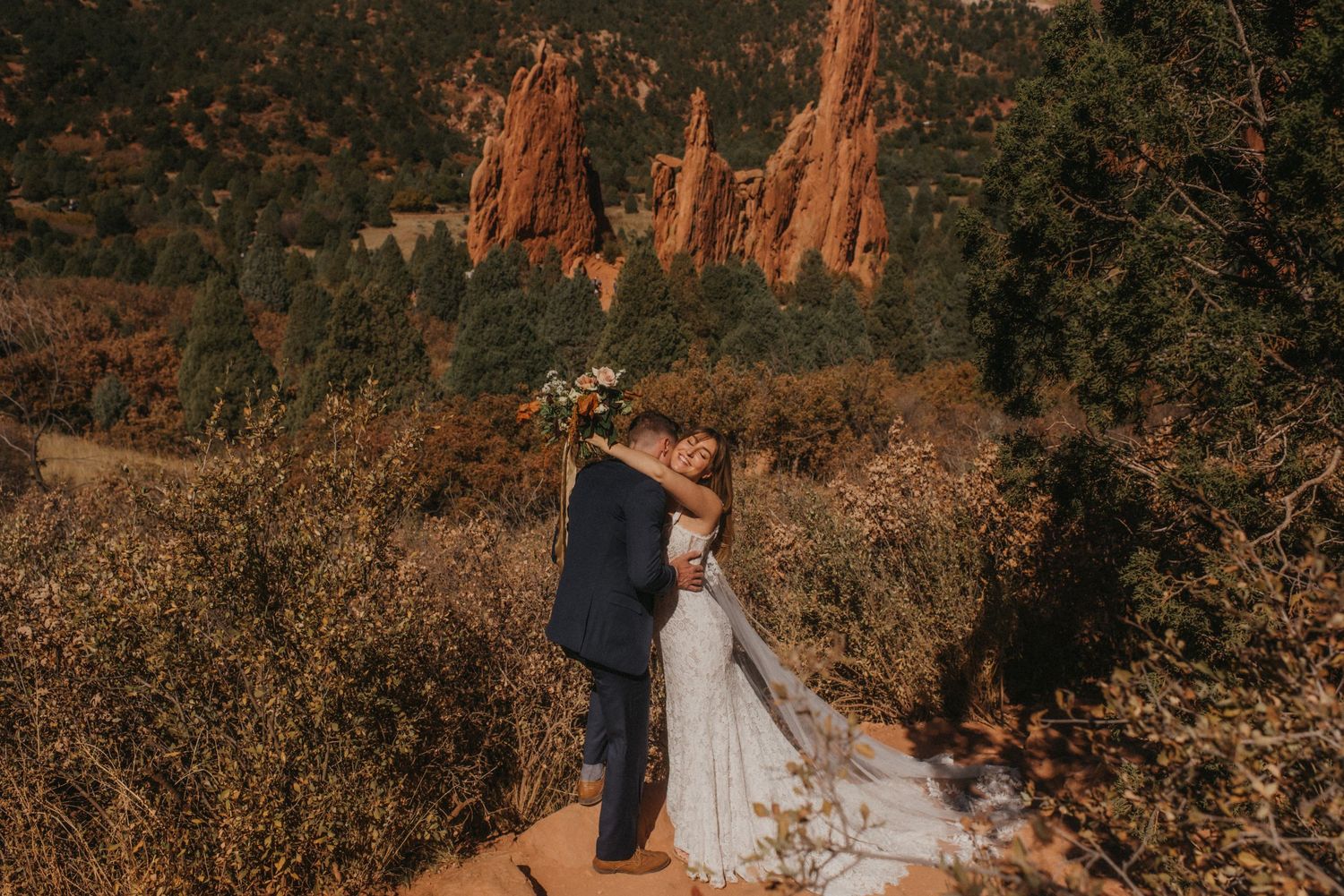 Couple in wedding attire embrace at sunset with red rock formations visible in Garden of the Gods, Colorado.