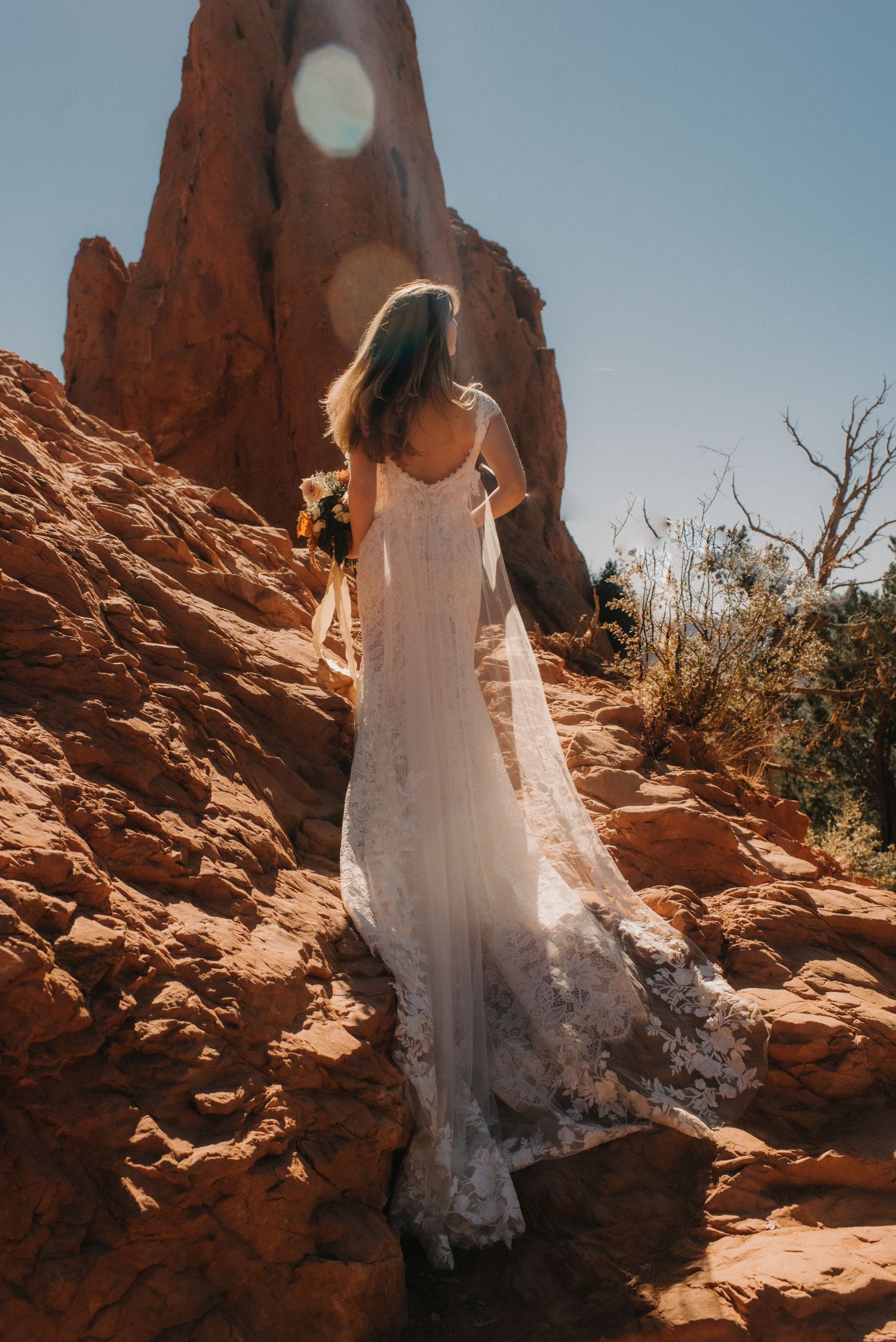 A bride in a white lace dress stands on red rock formations under bright sunlight.