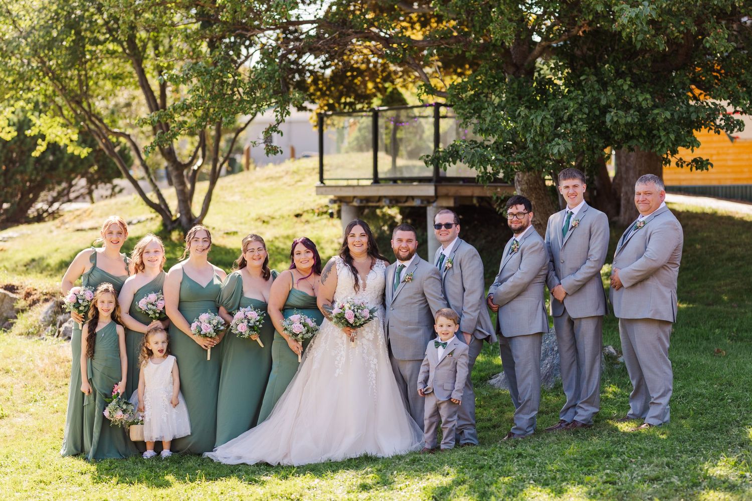 Wedding party in sage green dresses and gray suits poses together on a sunny hillside during outdoor celebration.