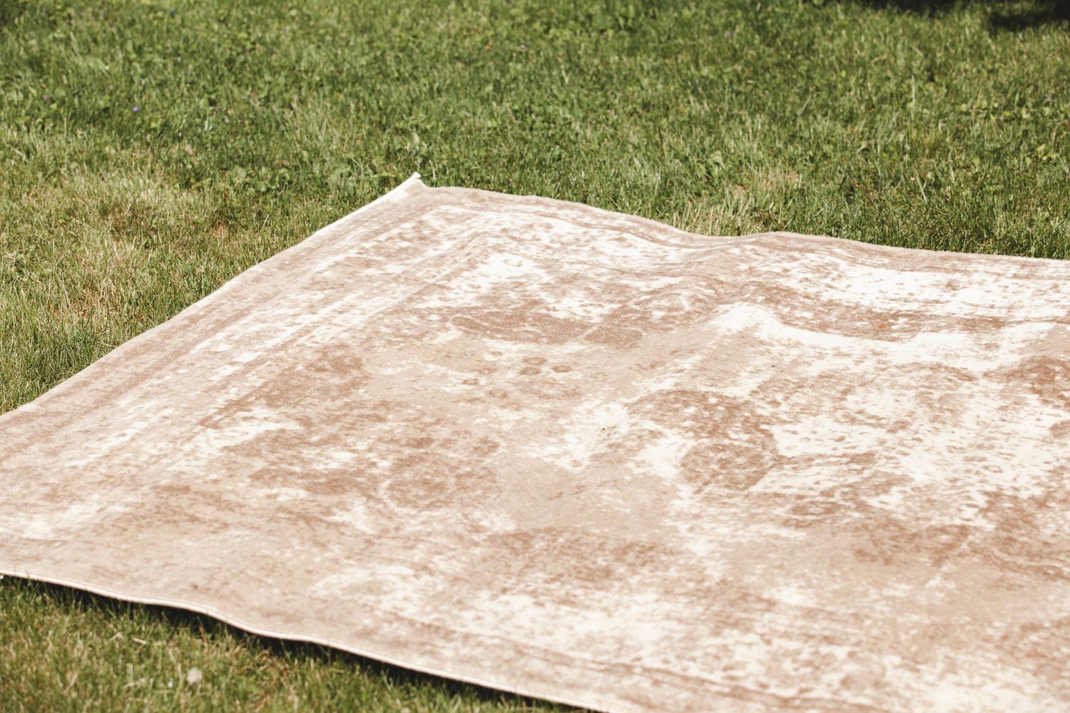 A well-worn beige area rug lies flat on green grass outdoors.