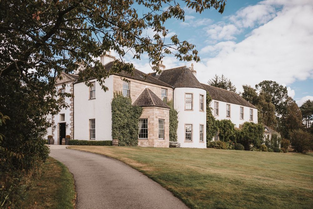 A stately white manor house with landscaped grounds and a curved driveway on a sunny day.
