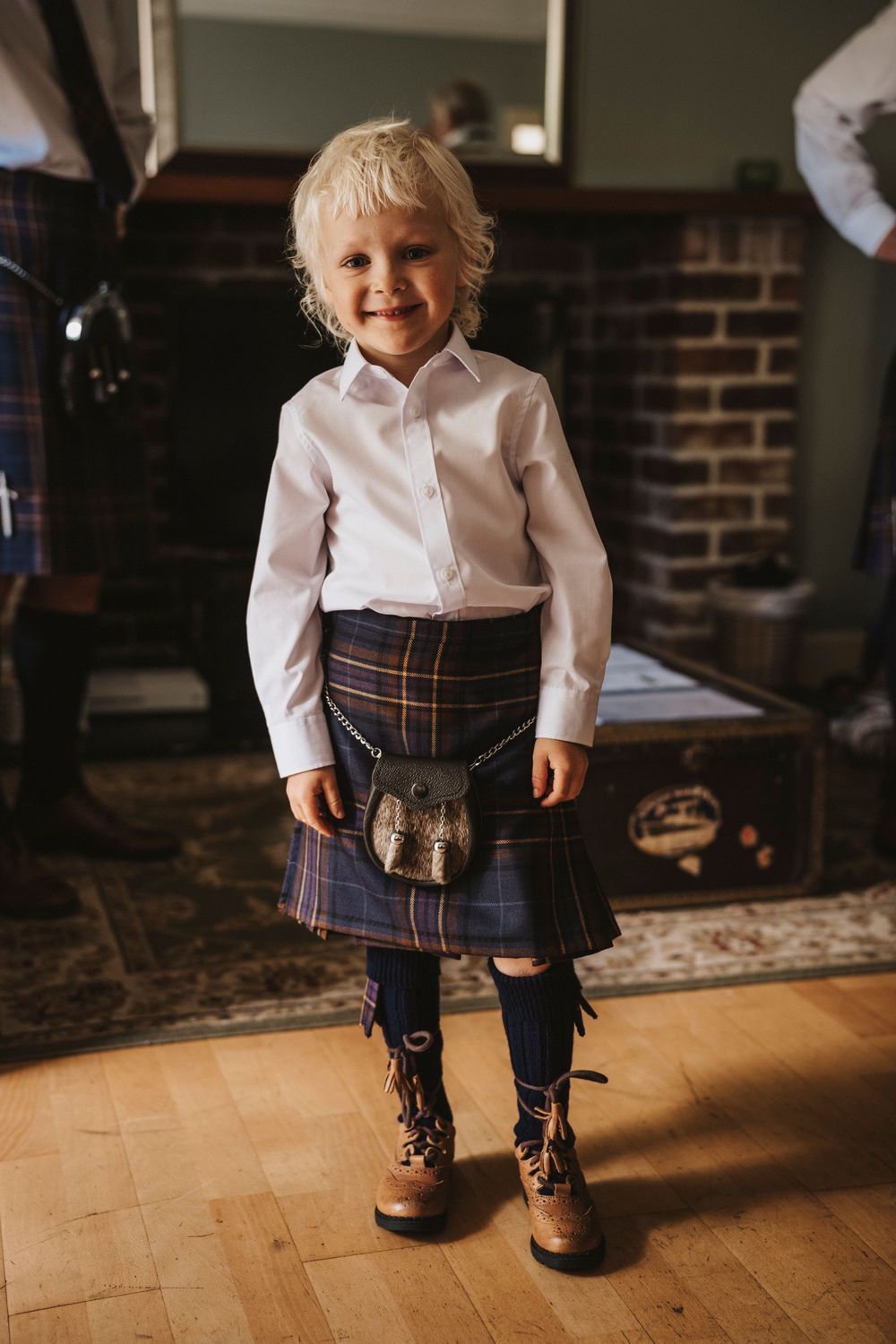 A child wearing a plaid pleated kilt and white shirt stands in different poses on a hardwood floor.