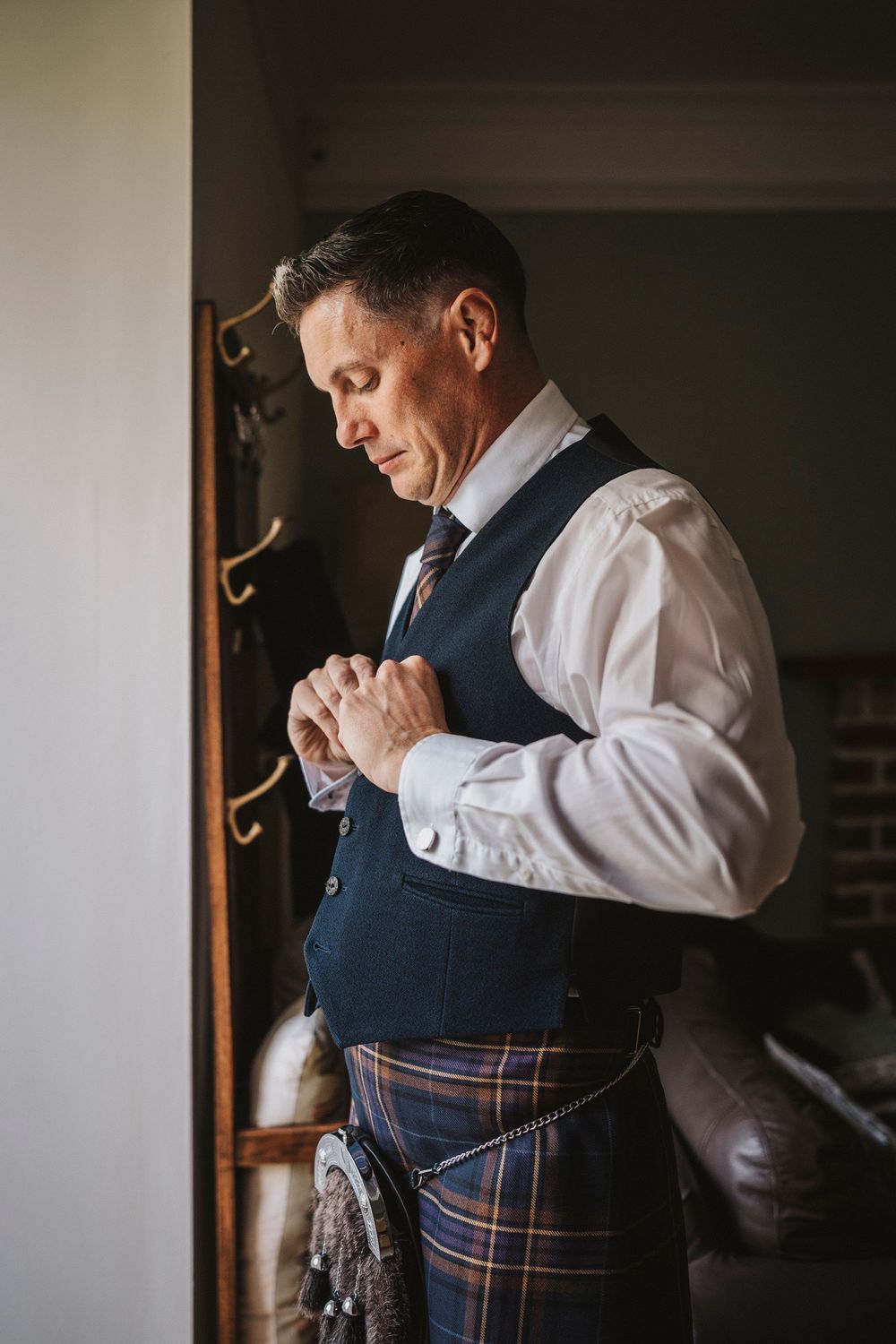 A groom in a navy vest and white shirt adjusts his tie while getting ready in natural window light.
