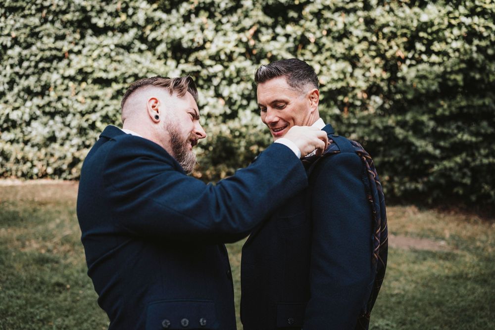 A romantic outdoor scene showing two people wearing navy suits embracing and smiling in front of leafy green foliage.