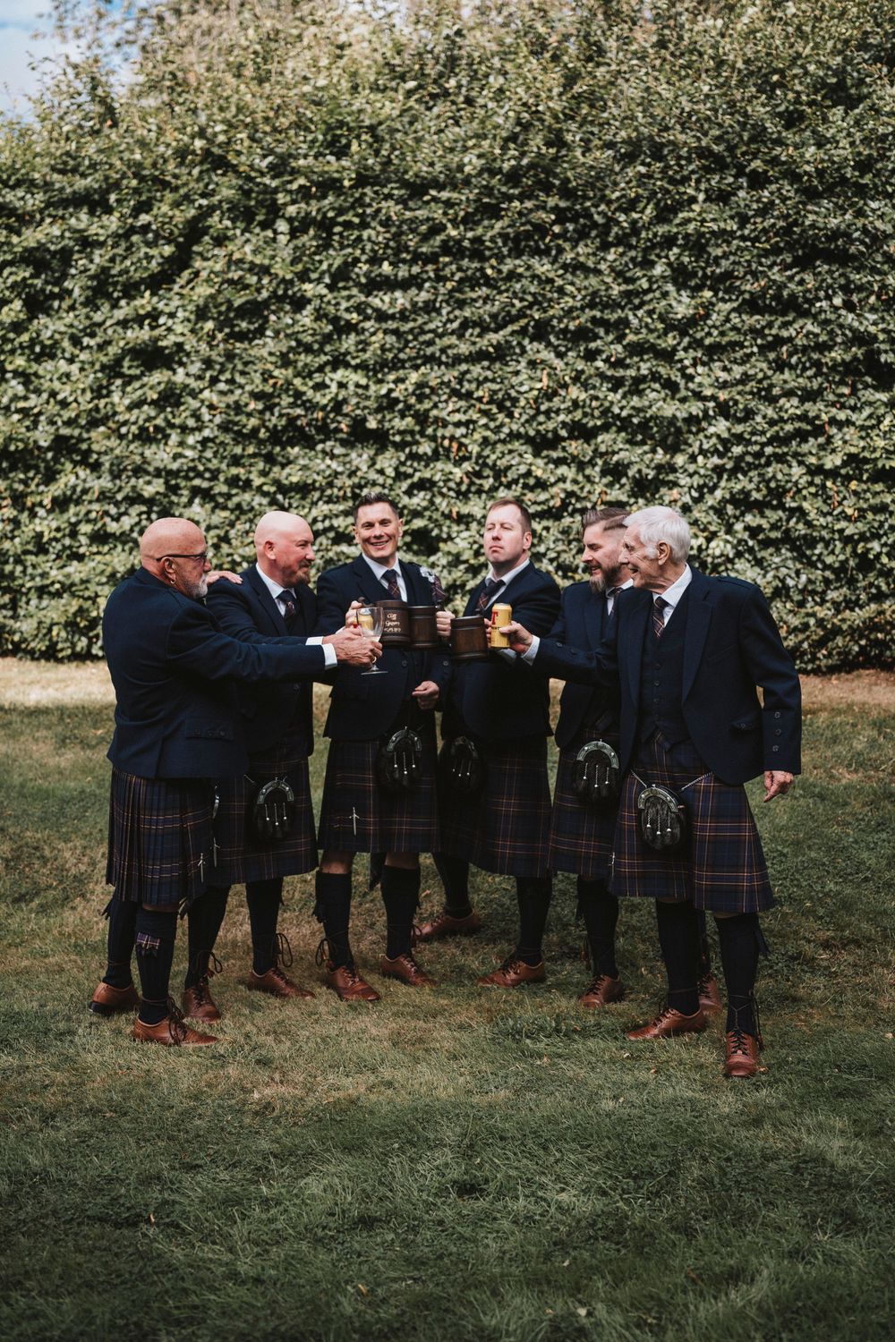 A group of men in traditional Scottish kilts stand together on grass raising drinks in a celebratory toast.