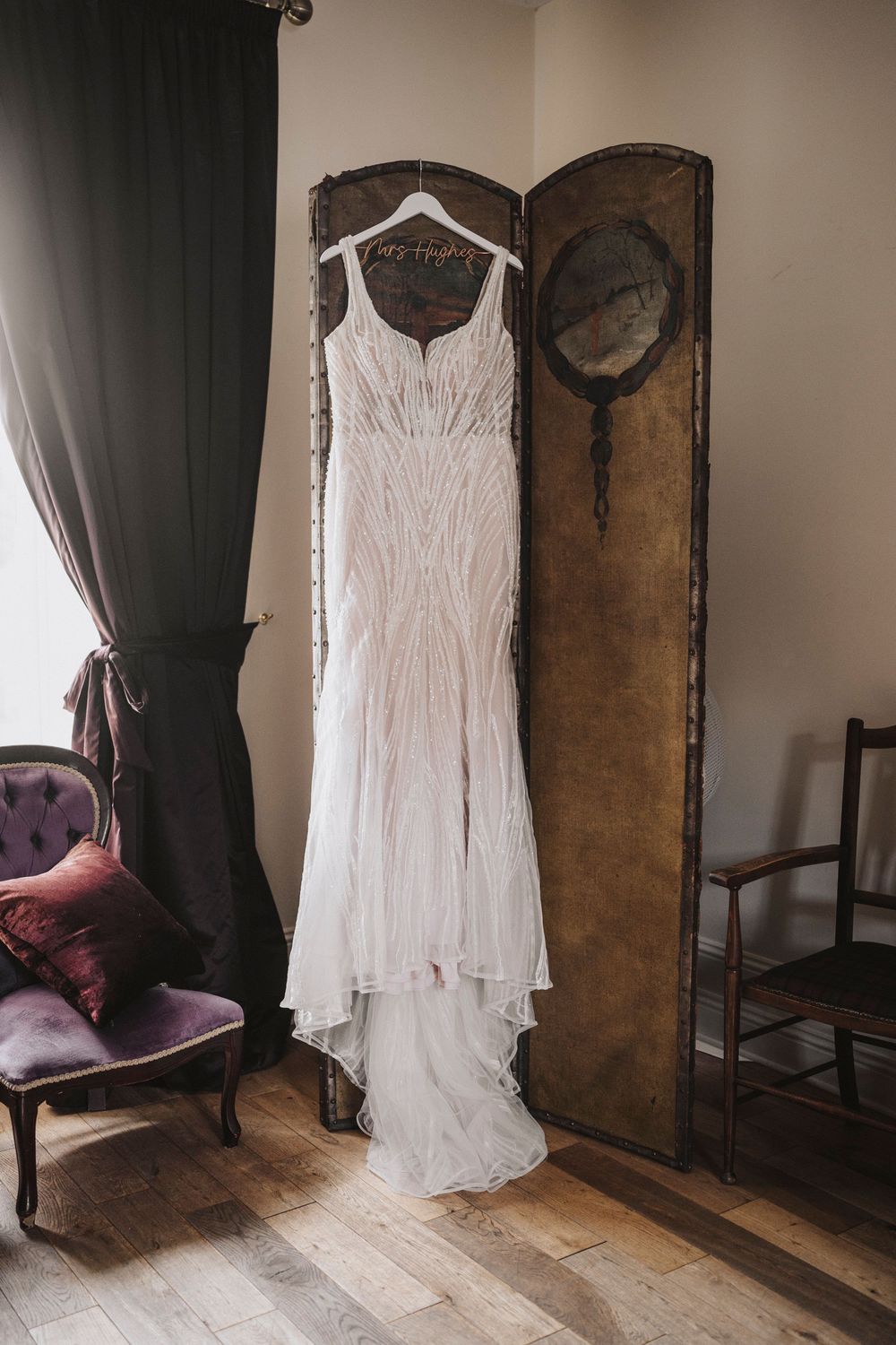 White lace wedding dress hanging elegantly against a dark wall with antique wooden furniture in view.