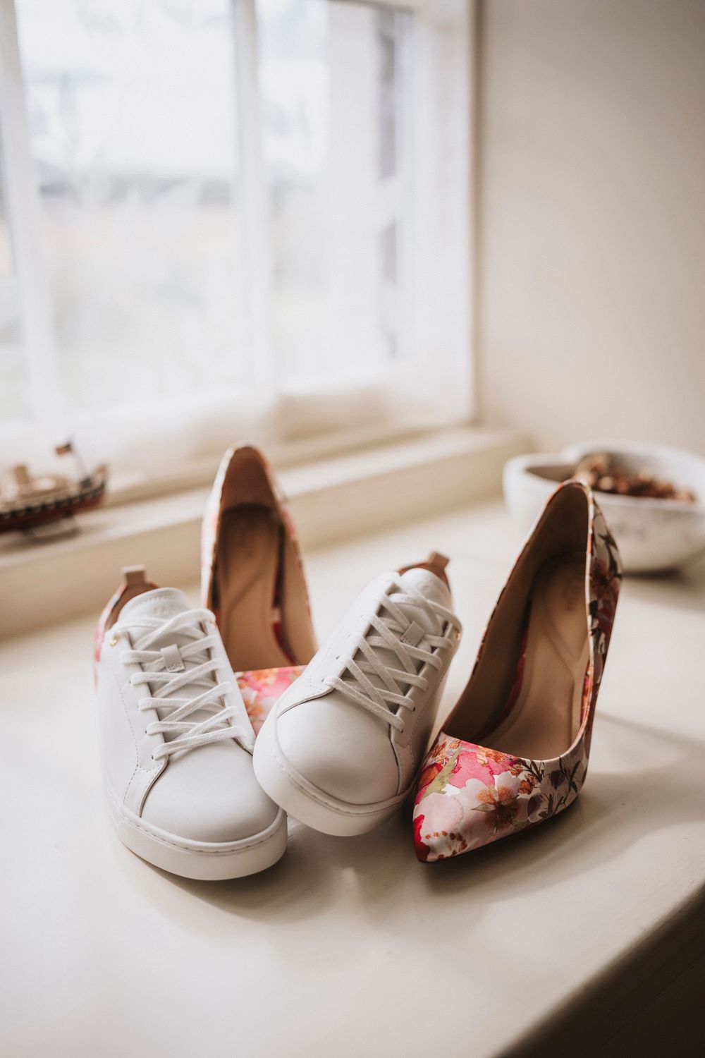 White sneakers and floral heels arranged on a windowsill, suggesting a bride's wedding day footwear choices.