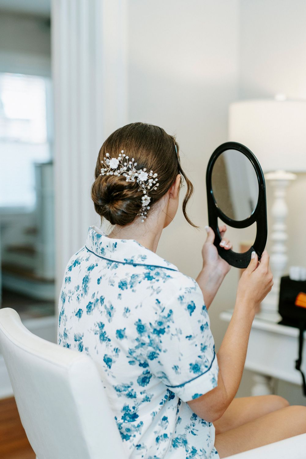 Person wearing blue floral pattern shirt doing hair styling and holding mirror in bright white room.