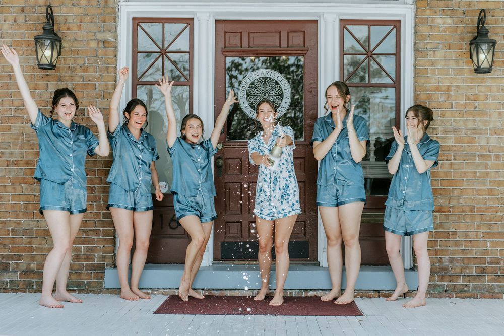 Bridal party wearing coordinated denim jackets poses in front of brick wall with lanterns.