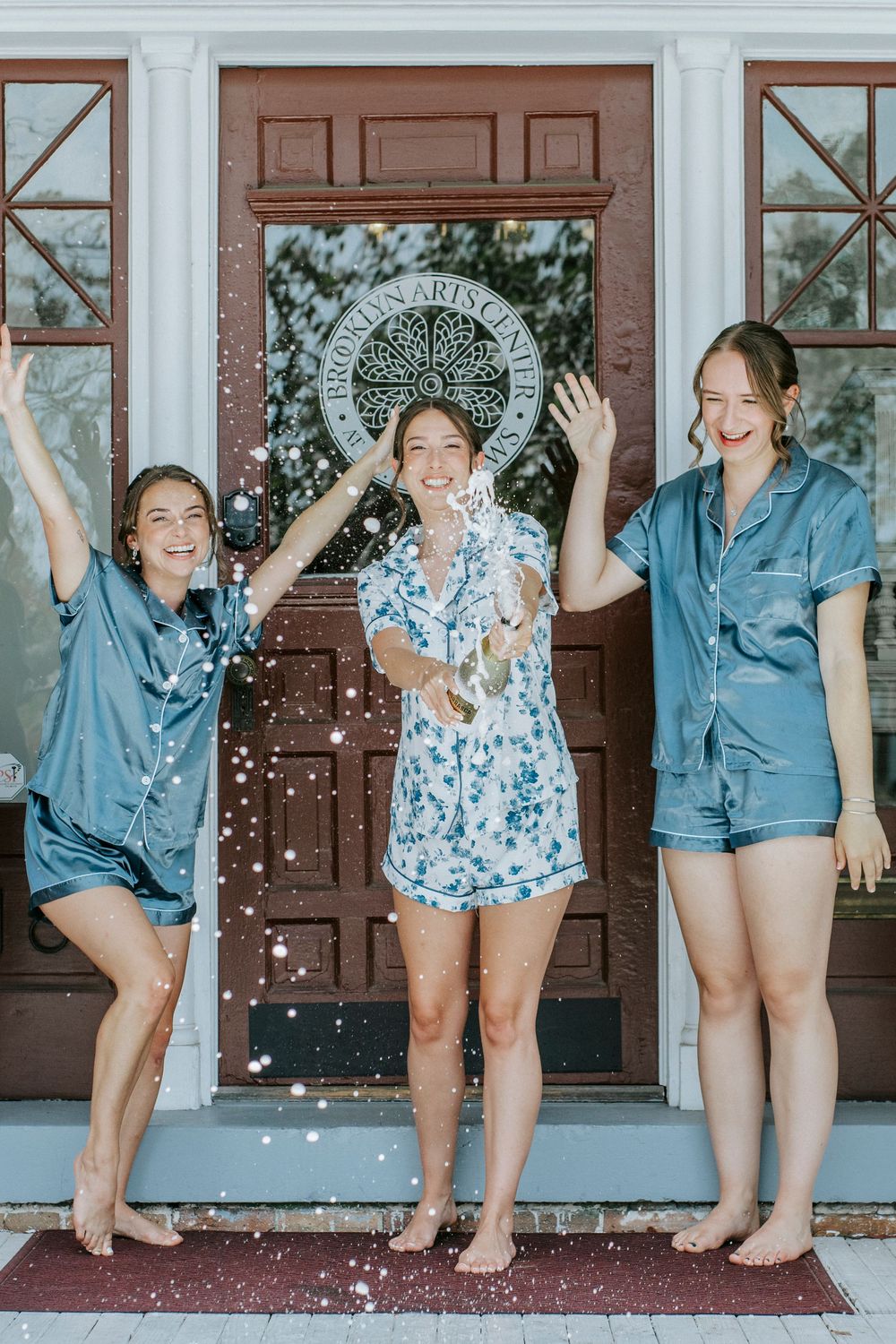 Three bridesmaids in blue pajamas celebrating joyfully on a front porch.