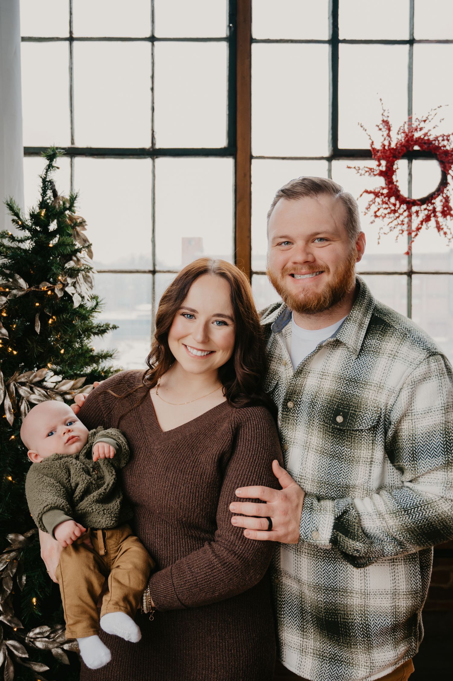 Smiling family with newborn baby posing near a Christmas tree by large industrial windows.