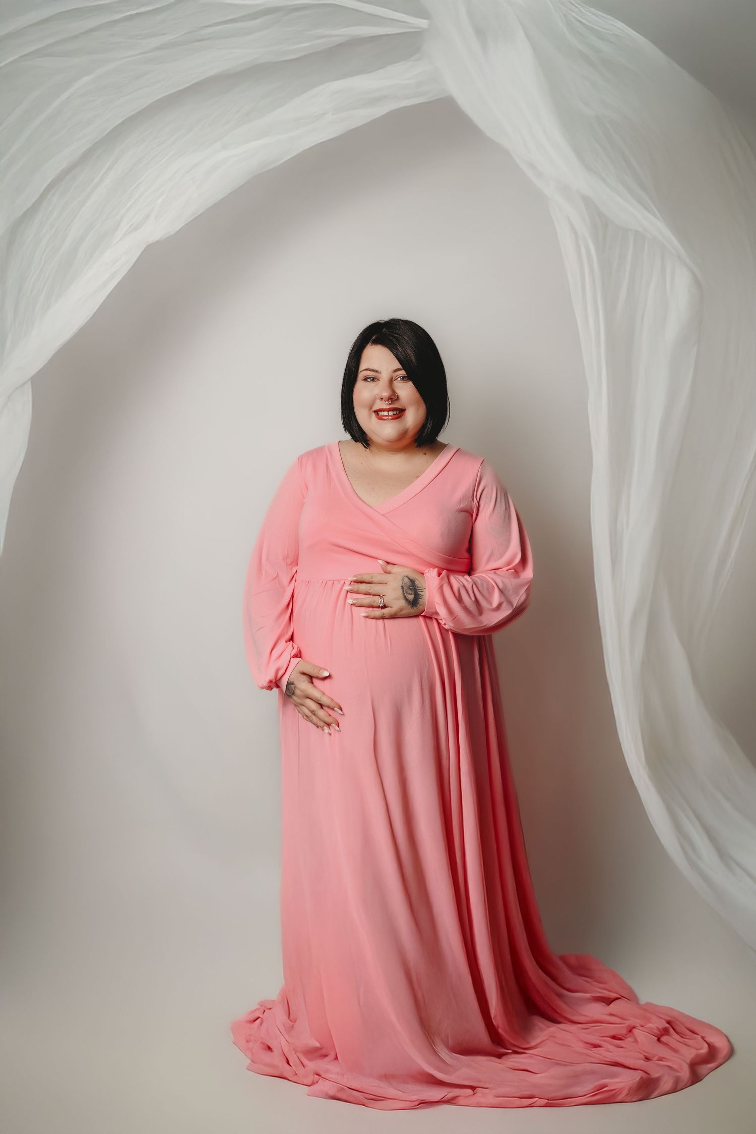Maternity portrait of woman in flowing pink dress against white fabric backdrop.
