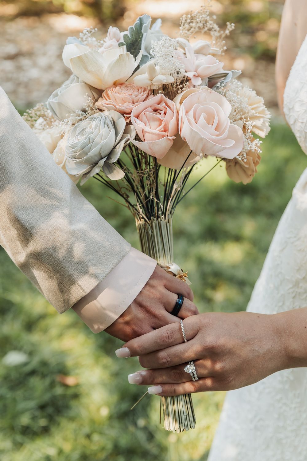 Hands holding a delicate bridal bouquet of blush pink and white roses with greenery against a soft outdoor background.