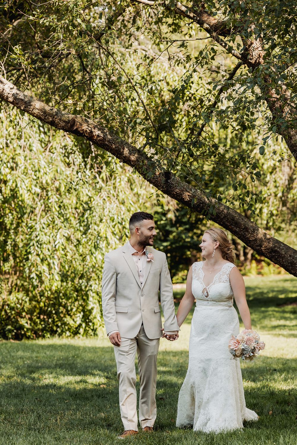 A romantic wedding couple shares a joyful moment together under a sunlit tree in their tan and white attire.
