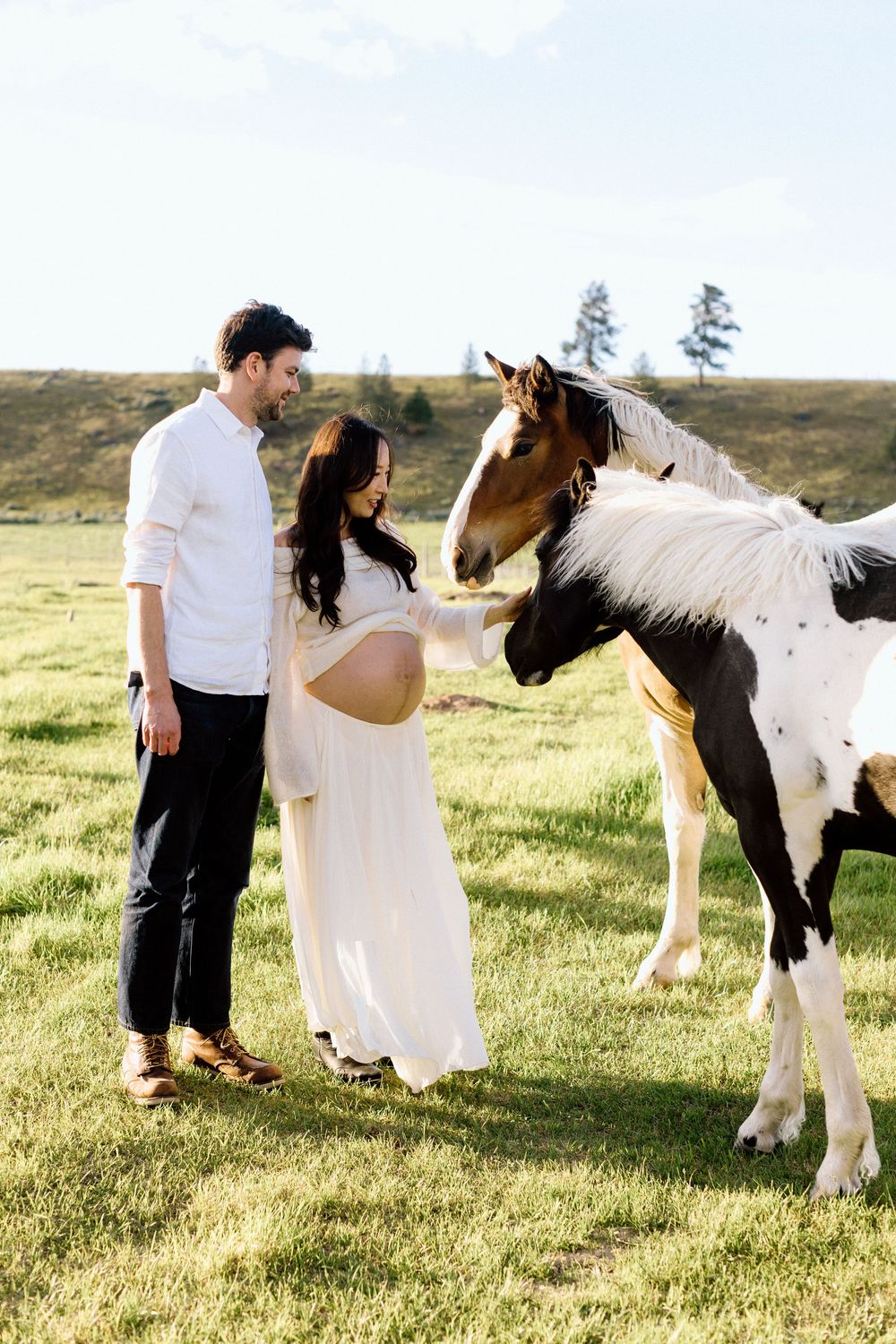 Pregnant couple in white attire tenderly interacting with a paint horse in a lush green field.