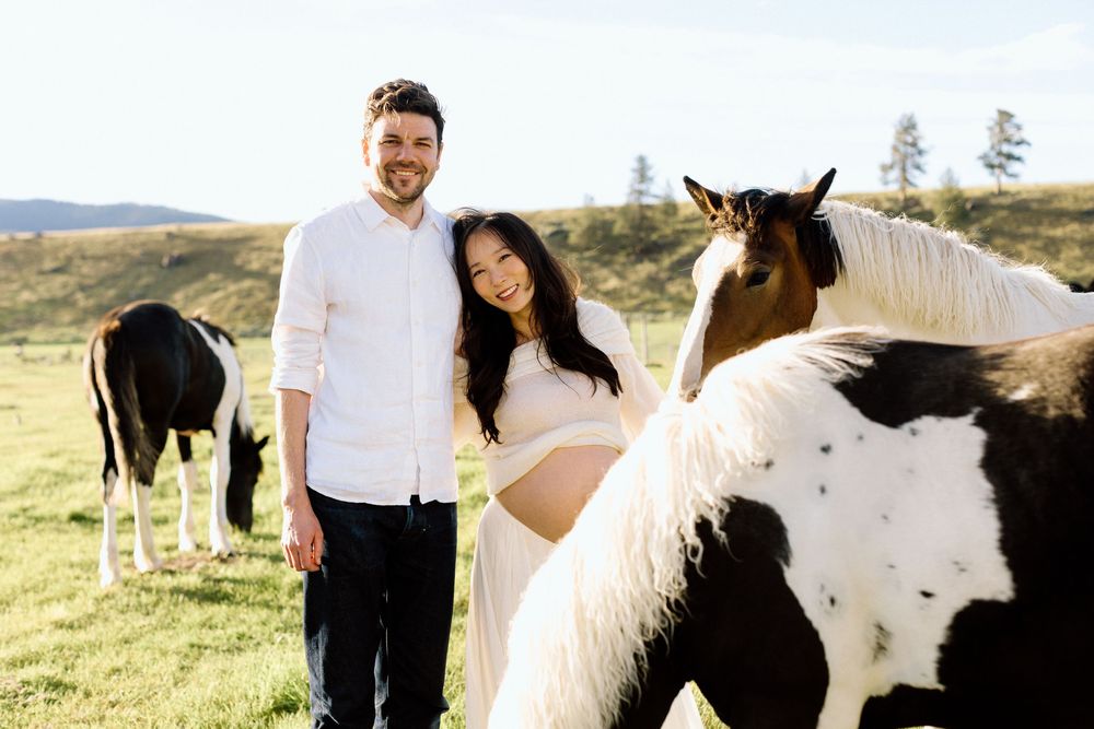 Pregnant couple smiling outdoors with black and white paint horses grazing on green pasture hills.