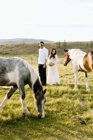 Pregnant couple in white outfits posing with grazing horses in a green field during a maternity photoshoot.