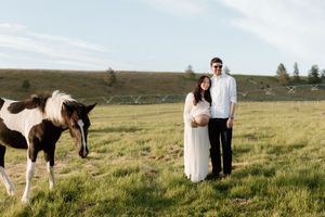Pregnant couple standing in green field with paint horse during outdoor maternity photo session.