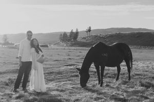Black and white photo of a couple posing in a field beside a grazing dark horse at sunset.