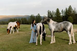 Pregnant woman in blue dress standing among paint horses in a scenic mountain meadow.