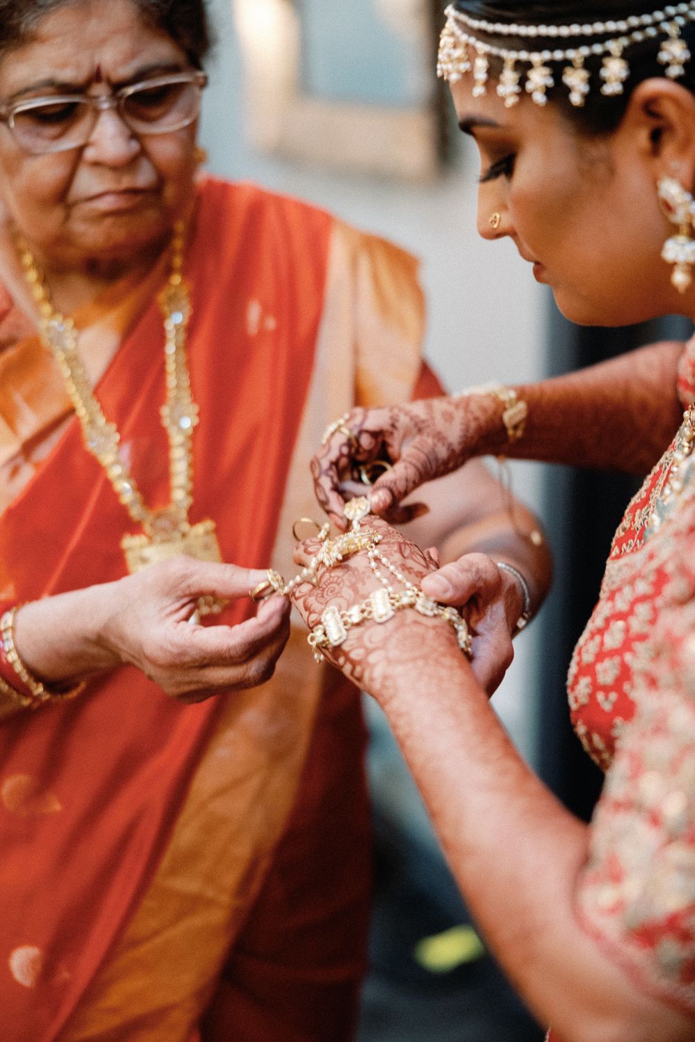 Elder woman in red saree helping bride with gold jewelry and henna-adorned hands during Indian wedding ceremony.