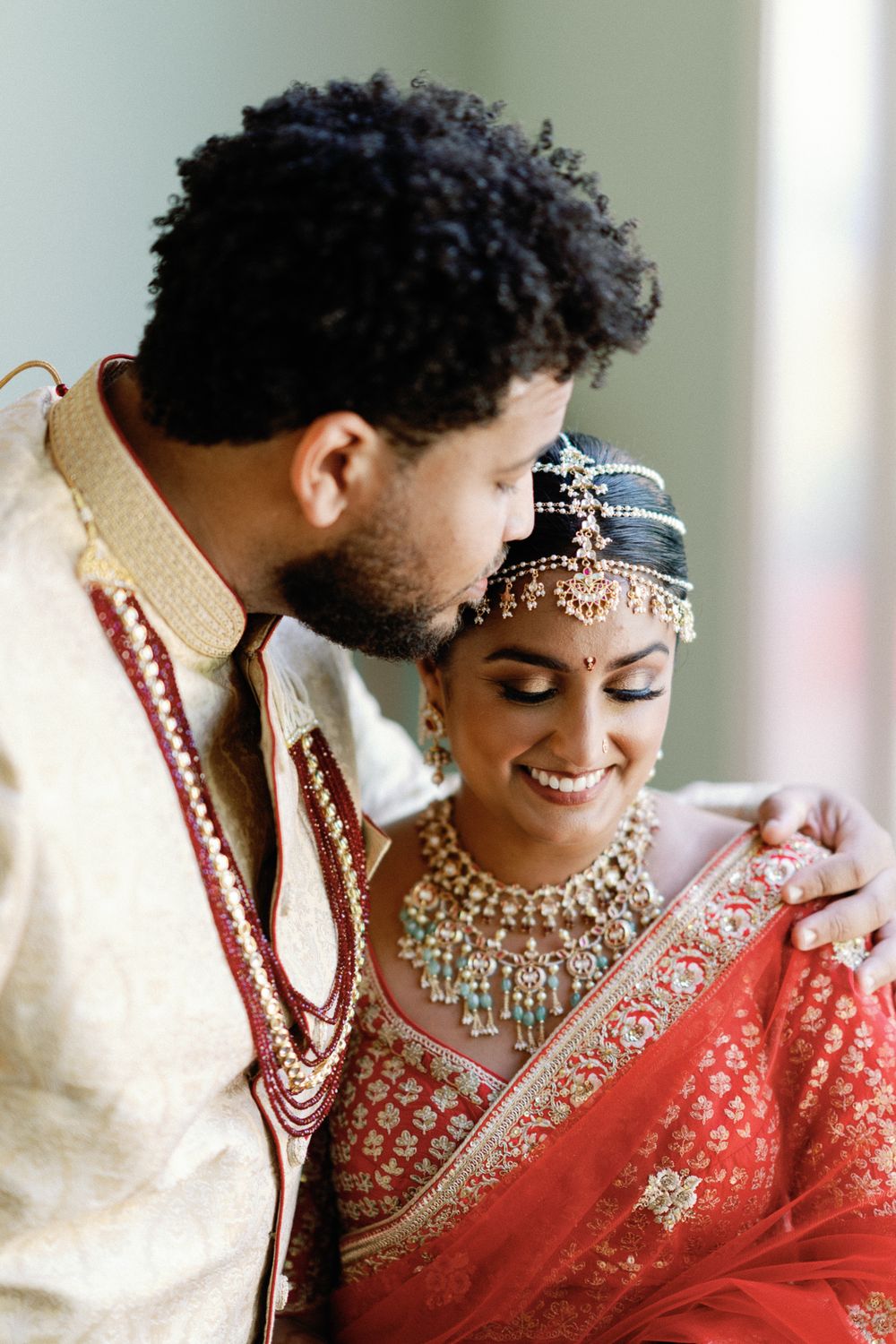Indian groom in gold sherwani kissing bride in red lehenga with kundan jewelry.