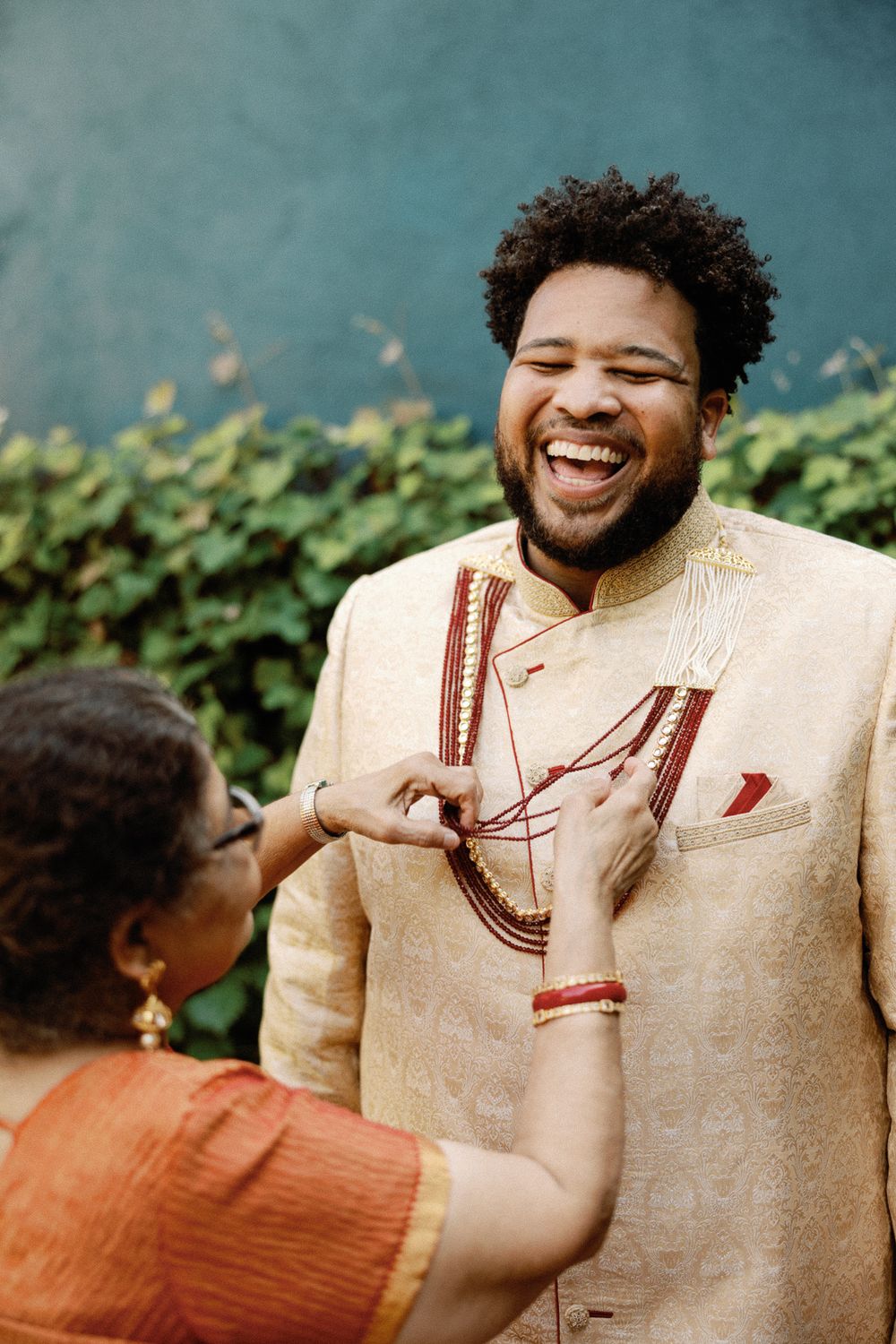 Joyful groom in gold sherwani laughing as a woman adjusts his wedding garlands outdoors.
